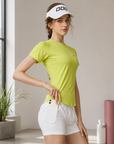 A woman wearing a bright lime green athletic t-shirt with a side pocket holding a phone, paired with white athletic shorts and a white visor, standing in a bright, modern indoor setting.