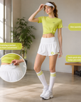 A woman modeling a bright yellow-green athletic crop top and white athletic skirt in a sunlit indoor room, with callout bubbles and an inset showing product features like a pocket and tech quick-dry fabric.